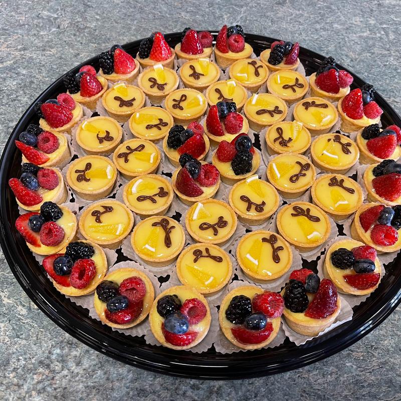 Large round platter of mini tartlettes, the centre filled with citrus tartlettes piped with a chocolate bow and the outer ring topped with strawberries, raspberries, blackberries and blueberries.
