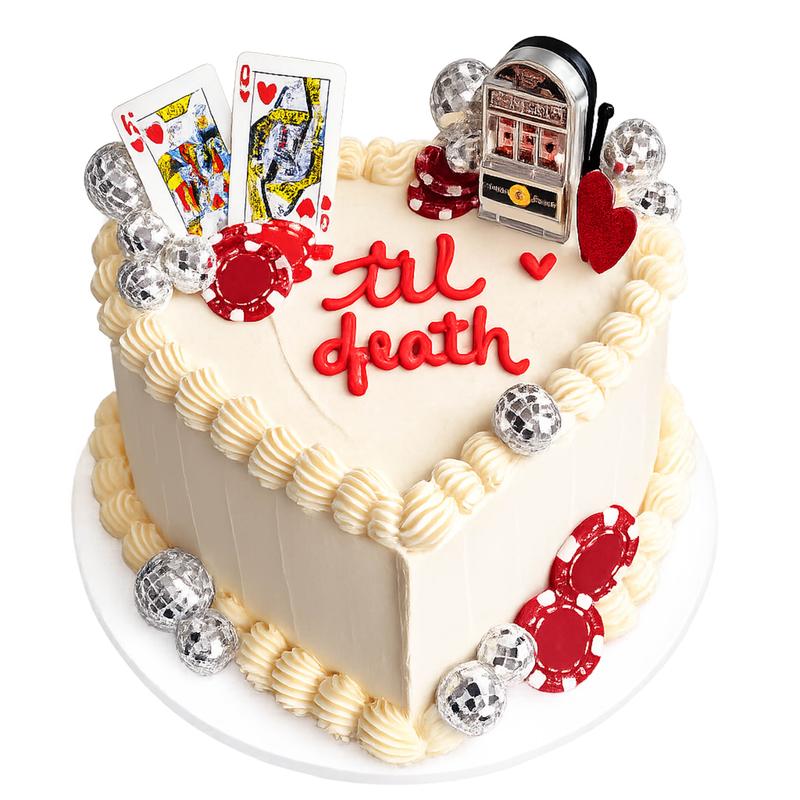 Heart-shaped ivory buttercream cake with piped shell borders and til death lettered in red, decorated with a king and queen of hearts, red poker chips, a slot-machine topper and mirrored disco balls.