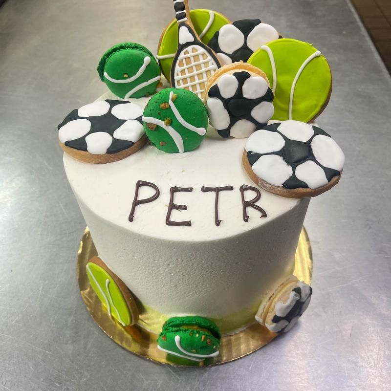 White buttercream cake lettered PETR in chocolate, crowned with iced cookies and macarons decorated as soccer balls, tennis balls and a tennis racquet.