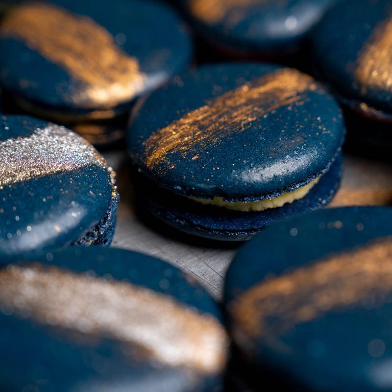 Close-up of custom macarons in deep navy shells brushed with broad strokes of gold and silver lustre, one turned to show its pale filling.
