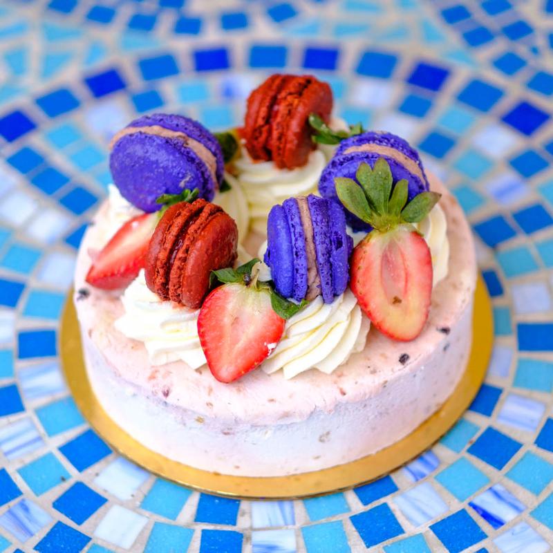 Fruits Des Bois cake: a pale berry mousse ringed with piped cream, fresh strawberries and purple and red macarons, on a gold board over a blue mosaic table.