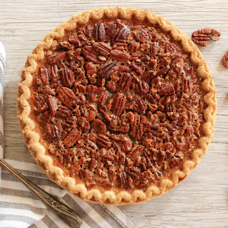 Pecan pie seen from above in a fluted crust, the glazed nuts packed edge to edge, with loose pecans and a vintage fork on the linen beside it.