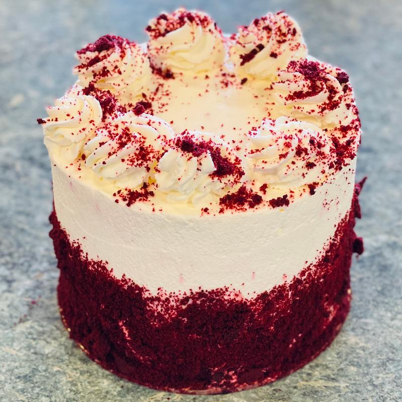 Red velvet cake finished in cream cheese frosting, the exposed lower band showing deep red crumb, with piped rosettes and red crumb scattered over the top.