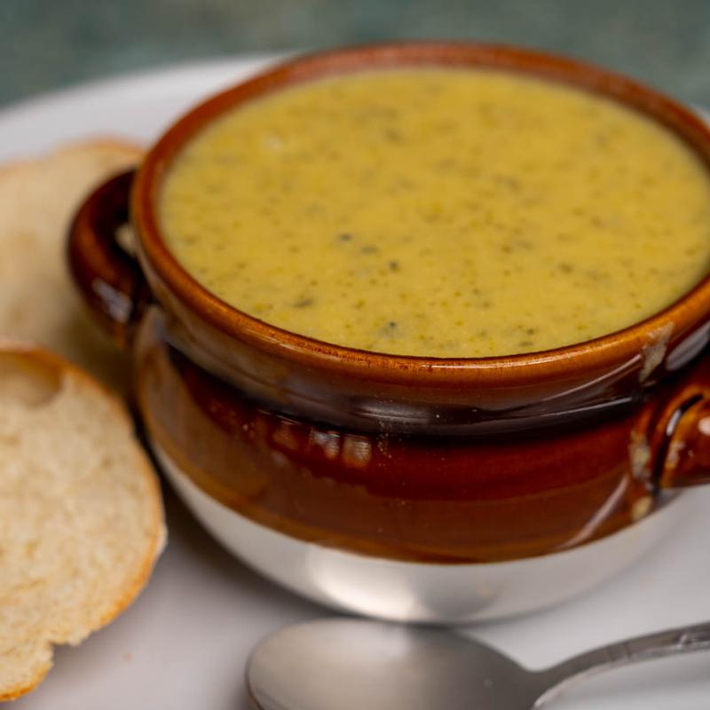 Close-up of a glazed brown crock of creamy broccoli soup with slices of baguette alongside.