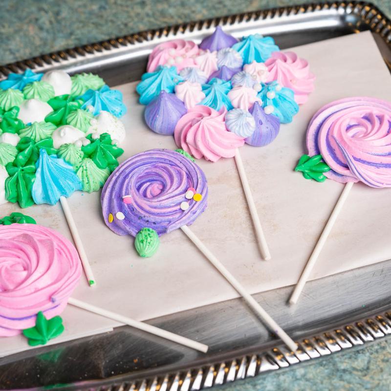 Meringue lollipops on a metal tray, piped as pink and purple swirls with green leaves, beside trays of small green, blue and white meringue kisses.