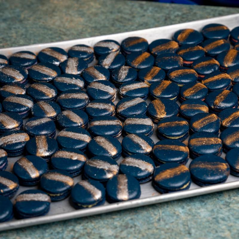 Baking tray filled with deep navy macaron shells, each brushed with a broad stripe of gold or silver lustre.