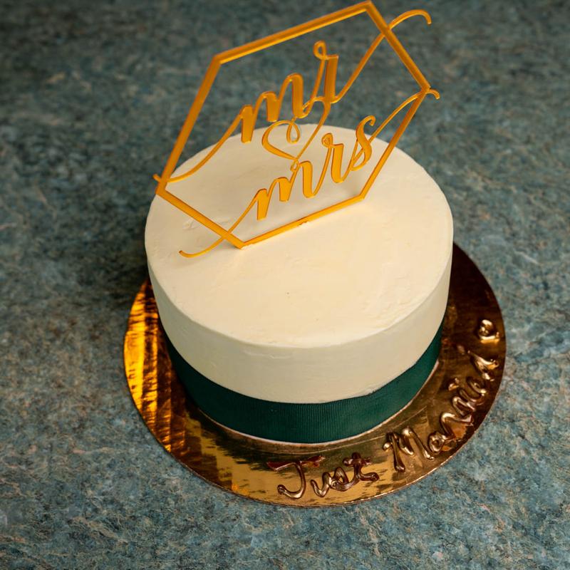 Single-tier ivory cake wrapped in a forest-green ribbon with a gold hexagon mr and mrs topper, on a gold board lettered Just Married.