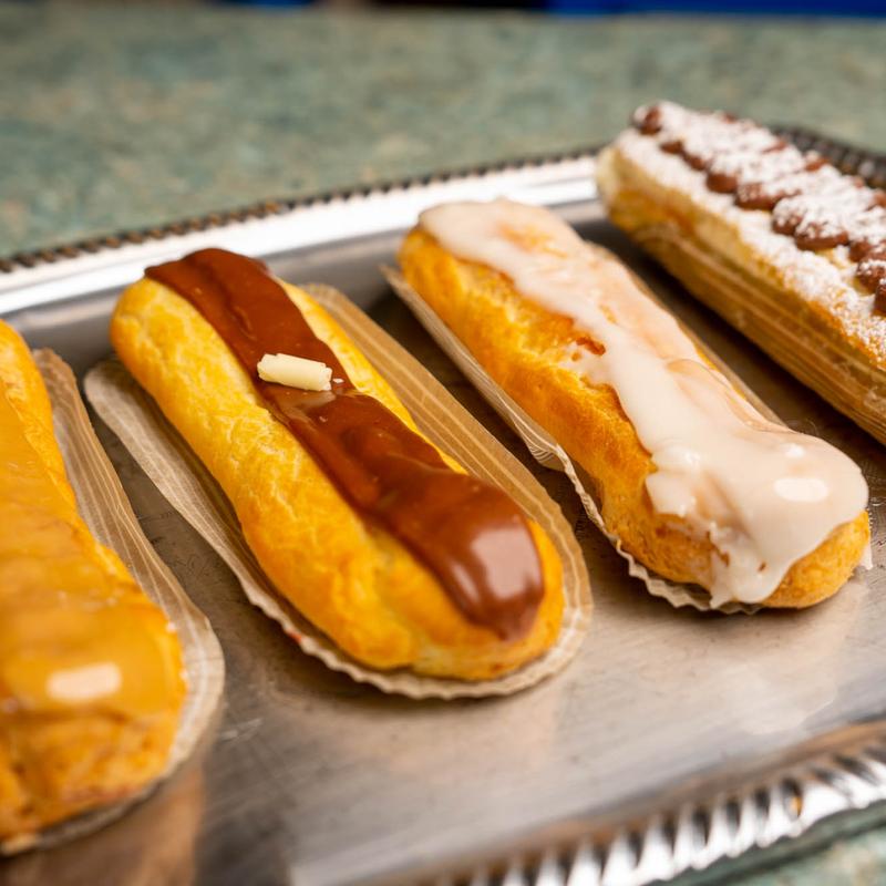 Eclairs on a metal tray, glazed in chocolate, vanilla fondant and coffee, with one at the back piped with cream and dusted with icing sugar.