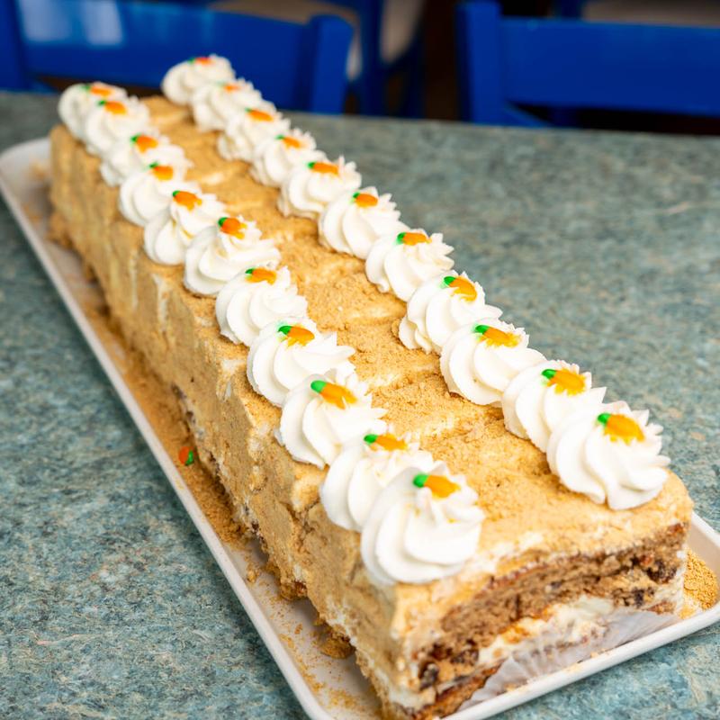 Long carrot loaf cake coated in cake crumb on a white platter, lined the full length with piped cream rosettes each finished with a tiny orange carrot.