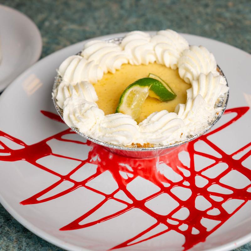 Individual citrus tart in a foil case, ringed with piped whipped cream and topped with a twist of lime, on a white plate decorated with a lattice of red fruit sauce.