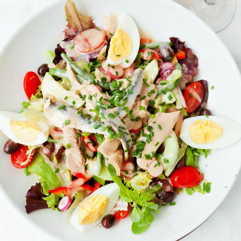 Composed salad in a white bowl: mixed leaves, green beans, cherry tomatoes, radish, olives, halved boiled eggs, flaked tuna and anchovy fillets, scattered with chives.