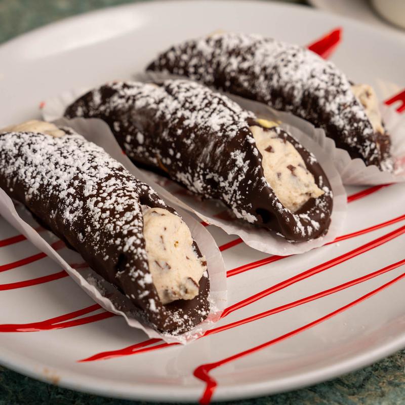 Three cannoli in chocolate-coated shells filled with chocolate-chip ricotta cream and dusted with icing sugar, on a plate striped with red fruit sauce.