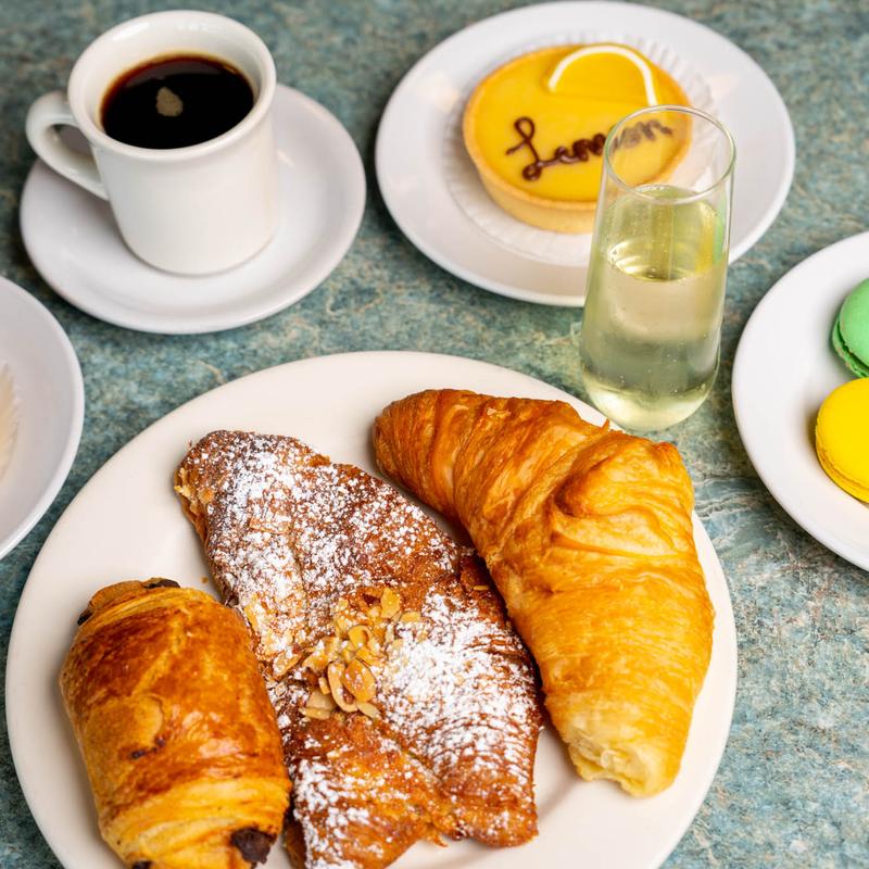 Cafe table set with black coffee, a citrus tartlette lettered in chocolate, a glass of sparkling wine, a plate of macarons and a plate of croissants, almond croissant and pain au chocolat.
