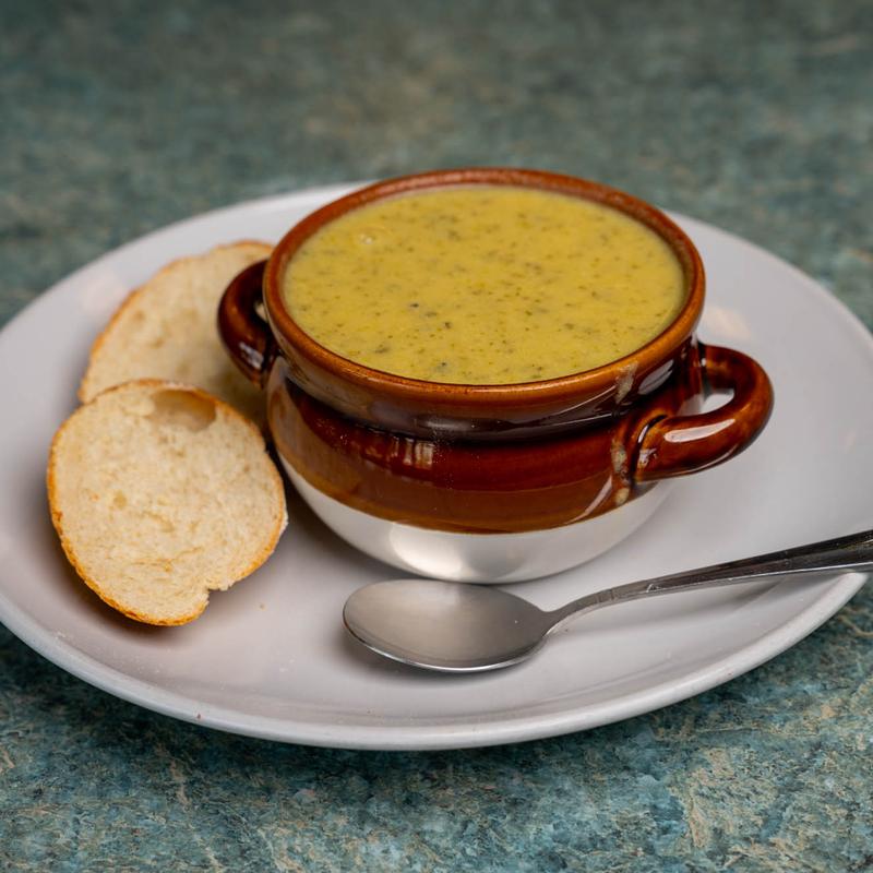 Glazed brown crock of creamy broccoli soup on a white plate with two slices of baguette and a spoon.