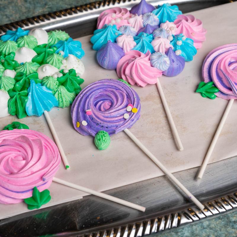 Meringue lollipops piped as pink and purple swirls on a metal tray, beside clusters of small green, blue and white meringue kisses.