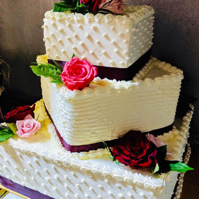 Three-tier square wedding cake in white buttercream, the tiers piped with quilting, ridges and shell borders, banded with plum ribbon and dressed with red and pink roses.