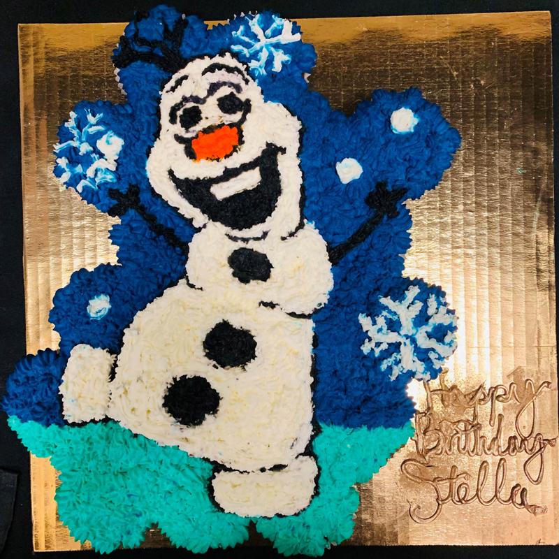 Square cake piped in buttercream as a smiling snowman on a blue snowflake background, with Happy Birthday Stella lettered on the gold board.
