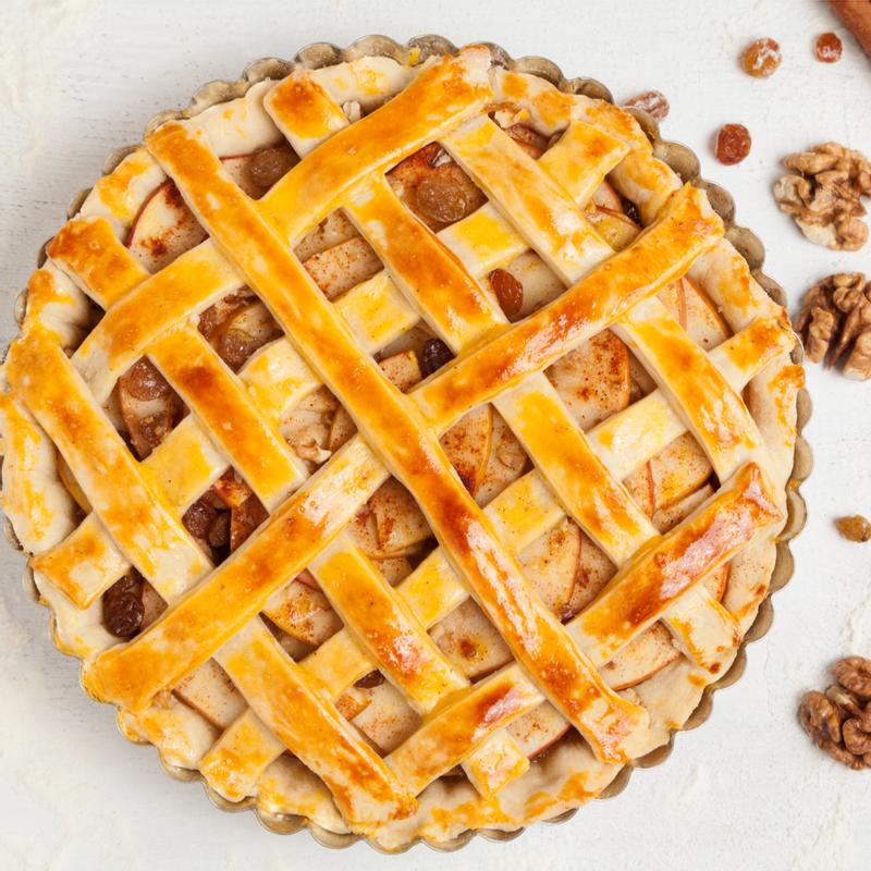 Lattice-topped apple pie seen from above in a fluted tin, the glazed pastry strips golden over spiced apple, with walnuts and raisins scattered on the table.