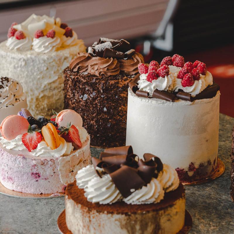 Group of round cakes on a stone counter: a raspberry-topped cream cake, a chocolate-crumb cake, a berry-and-macaron cake and a cocoa-dusted cake crowned with chocolate curls.
