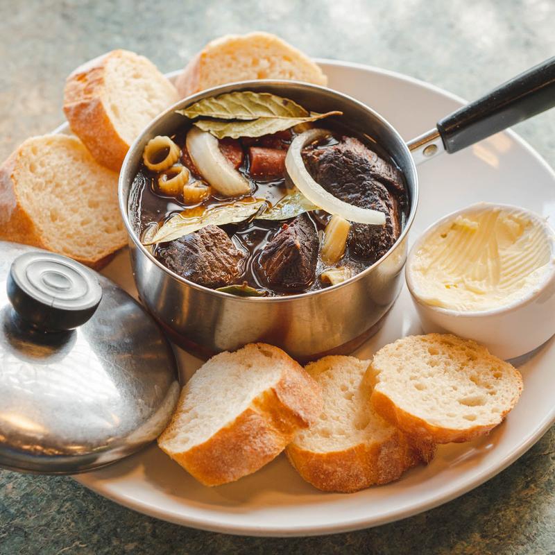 Beef stew served in a small steel pot with onion, bay leaves and pasta, on a plate with sliced baguette, a lidded dish and a ramekin of curled butter.