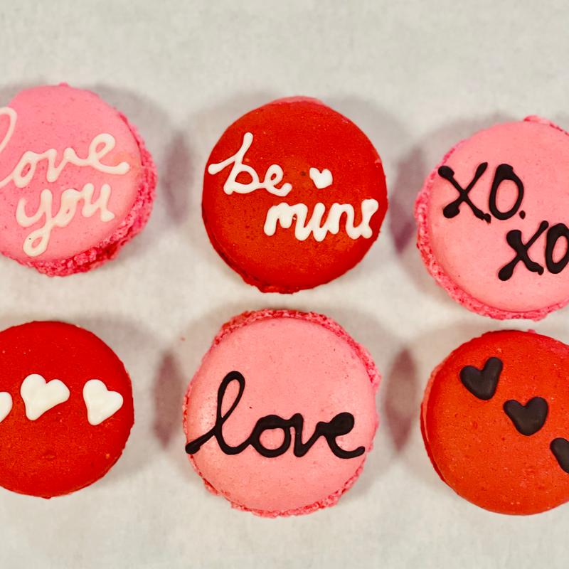 Six pink and red macarons piped with love you, be mine, love and xoxo in chocolate and icing, two of them dotted with small icing hearts.