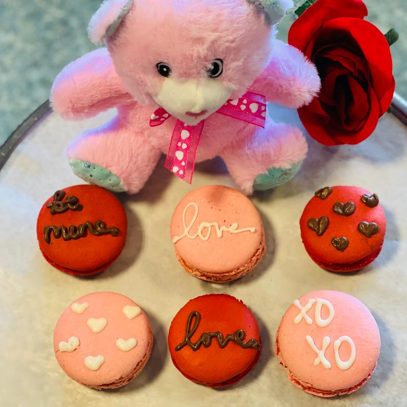Six pink and red macarons piped with be mine, love and xoxo in chocolate and icing, arranged in front of a small pink teddy bear and a red rose.