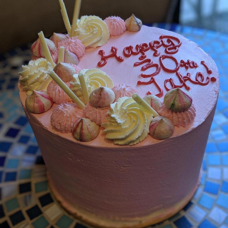 Pink buttercream cake lettered Happy 30th Jake in red icing, crowned with cream and pink meringue kisses, piped rosettes and white chocolate batons.