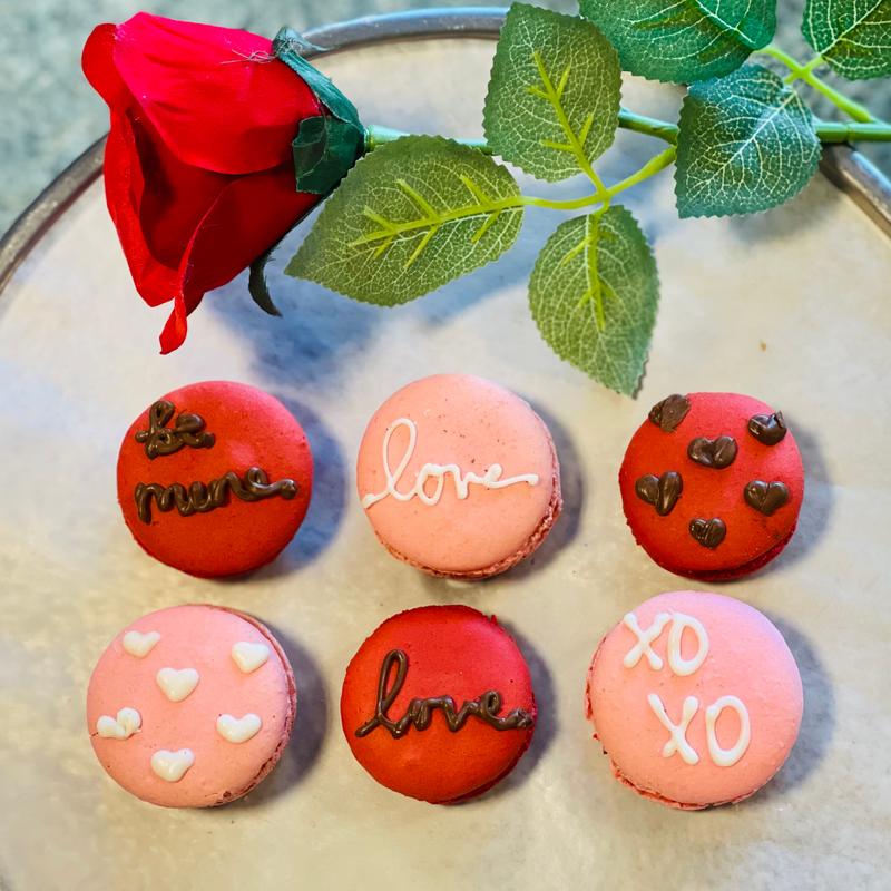 Six pink and red macarons piped with be mine, love and xoxo in chocolate and icing, arranged on a marble stand beside a red rose.