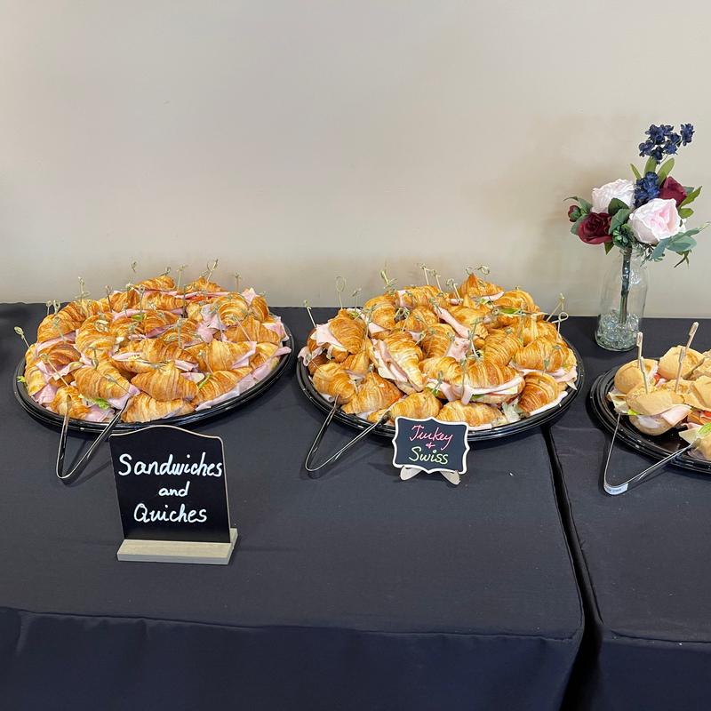 Catering table with two black platters of mini croissant sandwiches on cocktail picks, chalkboard signs reading Sandwiches and Quiches and Turkey and Swiss, and a jar of roses beside them.