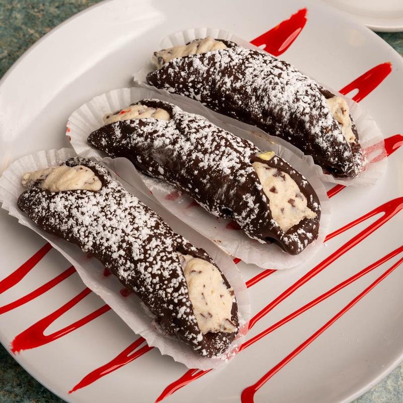 Three cannoli in chocolate-coated shells filled with ricotta cream and dusted with icing sugar, on a white plate drizzled with red fruit sauce.
