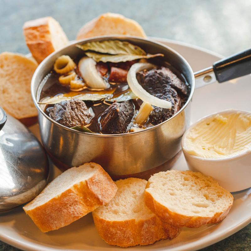 Close-up of beef stew in a steel pot with onion and bay leaves, surrounded by slices of baguette and a ramekin of curled butter.