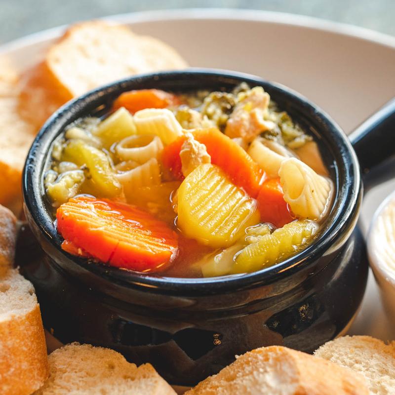 Black crock of vegetable and pasta soup with carrot, celery, courgette and beans, ringed with slices of baguette.