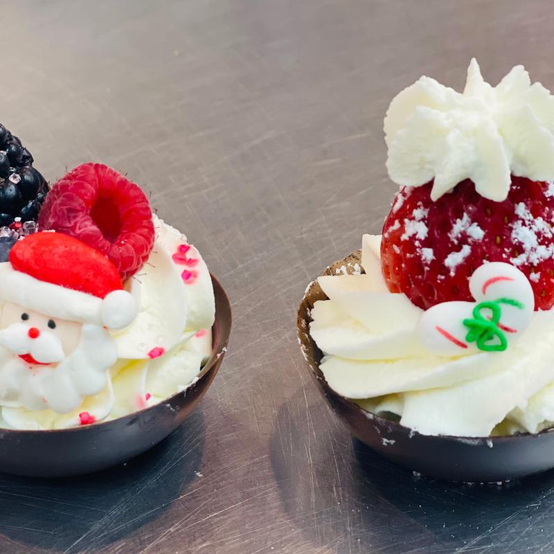 Two dark chocolate cups filled with piped cream, one topped with a sugar Santa, berries and sprinkles, the other with a strawberry and a sugar candy cane.