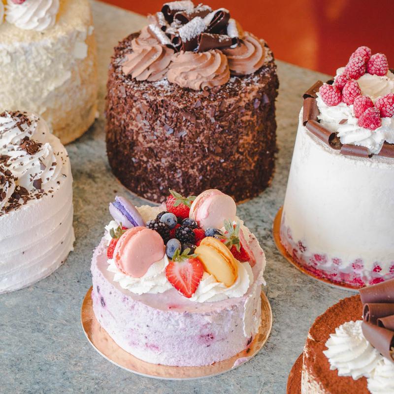 Round cakes on a stone counter: a coconut cream cake, a chocolate-crumb cake with chocolate curls, a raspberry cake, a berry-and-macaron cake and a cocoa-dusted cake.
