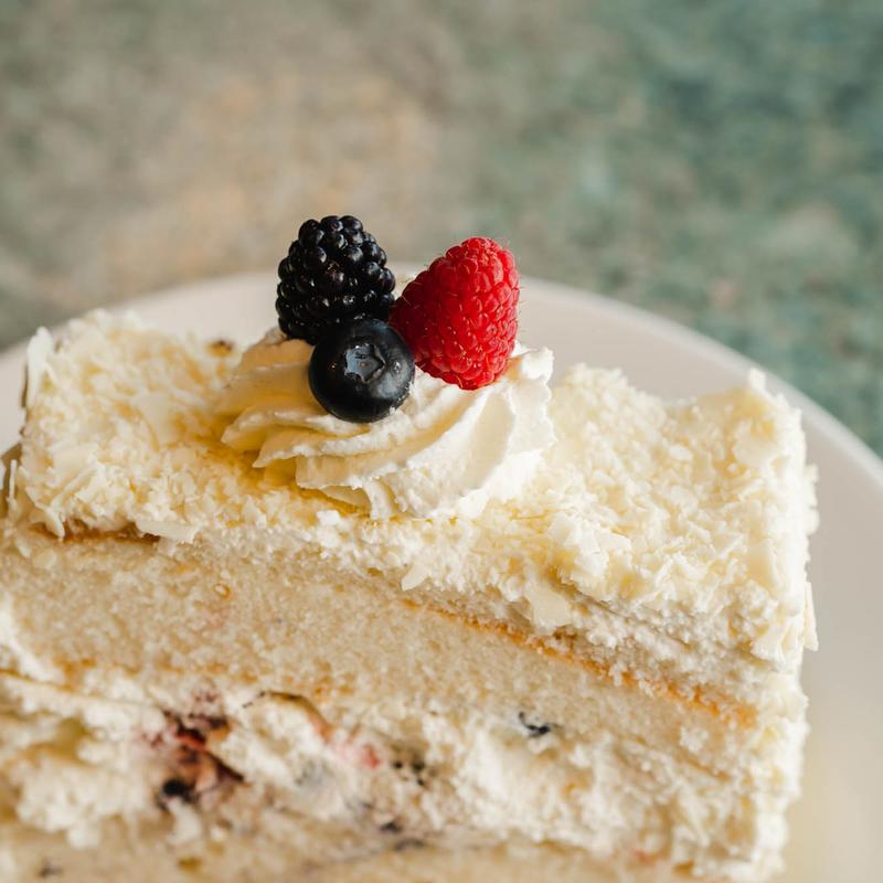Slice of white layer cake with cream between the sponges, the sides pressed with white chocolate shavings and the top finished with a piped rosette, a blackberry, a raspberry and a blueberry.