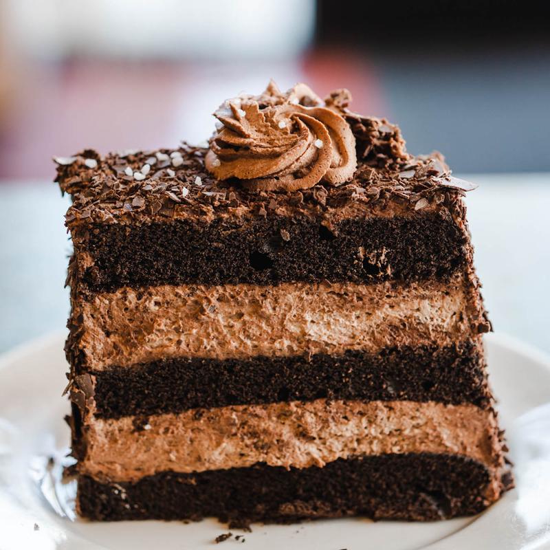 Tall slice of chocolate layer cake showing four dark sponge layers alternating with chocolate mousse, the sides pressed with chocolate shavings and the top finished with a piped rosette.