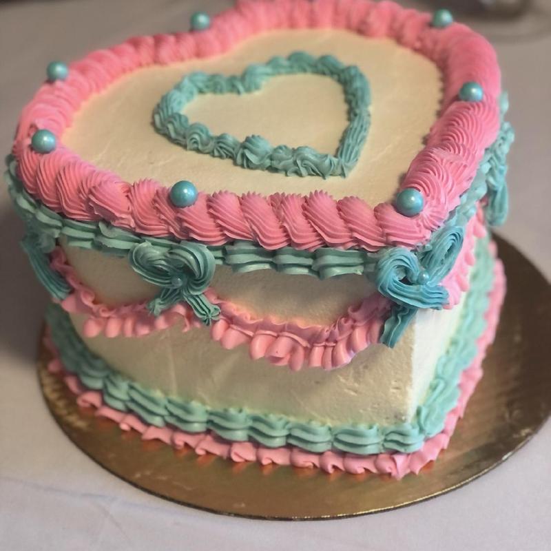 Heart-shaped cake in ivory buttercream with vintage piping: pink ruffled borders, turquoise rosettes and bows, blue sugar pearls and a piped turquoise heart on top.