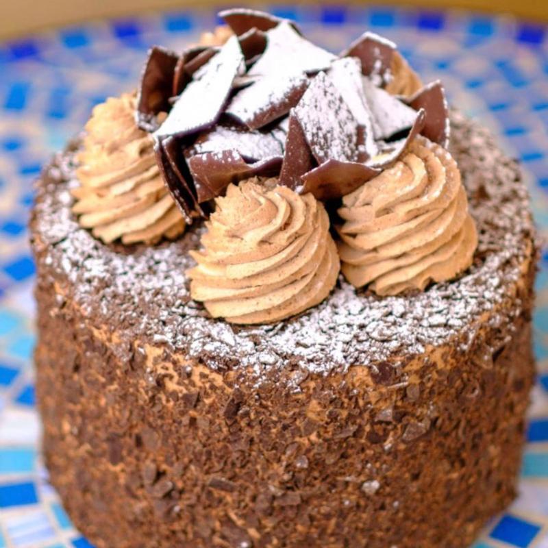 Truffle cake with sides coated in dark chocolate flakes, topped with piped chocolate cream rosettes, a pile of chocolate shards and a dusting of icing sugar.