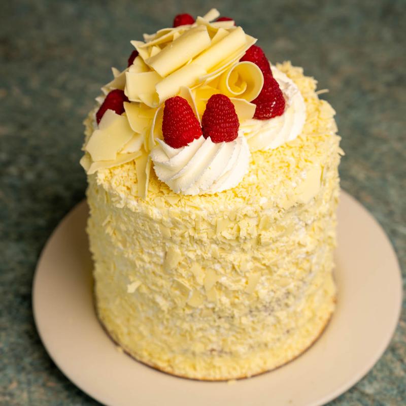 White raspberry cake covered in white chocolate flakes and crowned with white chocolate curls, piped cream and fresh raspberries.
