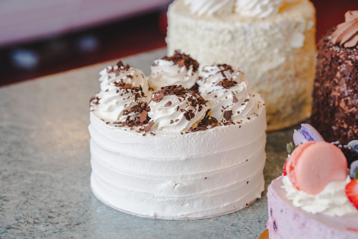 Gluten-free cake on the bakery counter, finished in smooth white cream and topped with piped rosettes and chocolate shavings, with other cakes behind it.