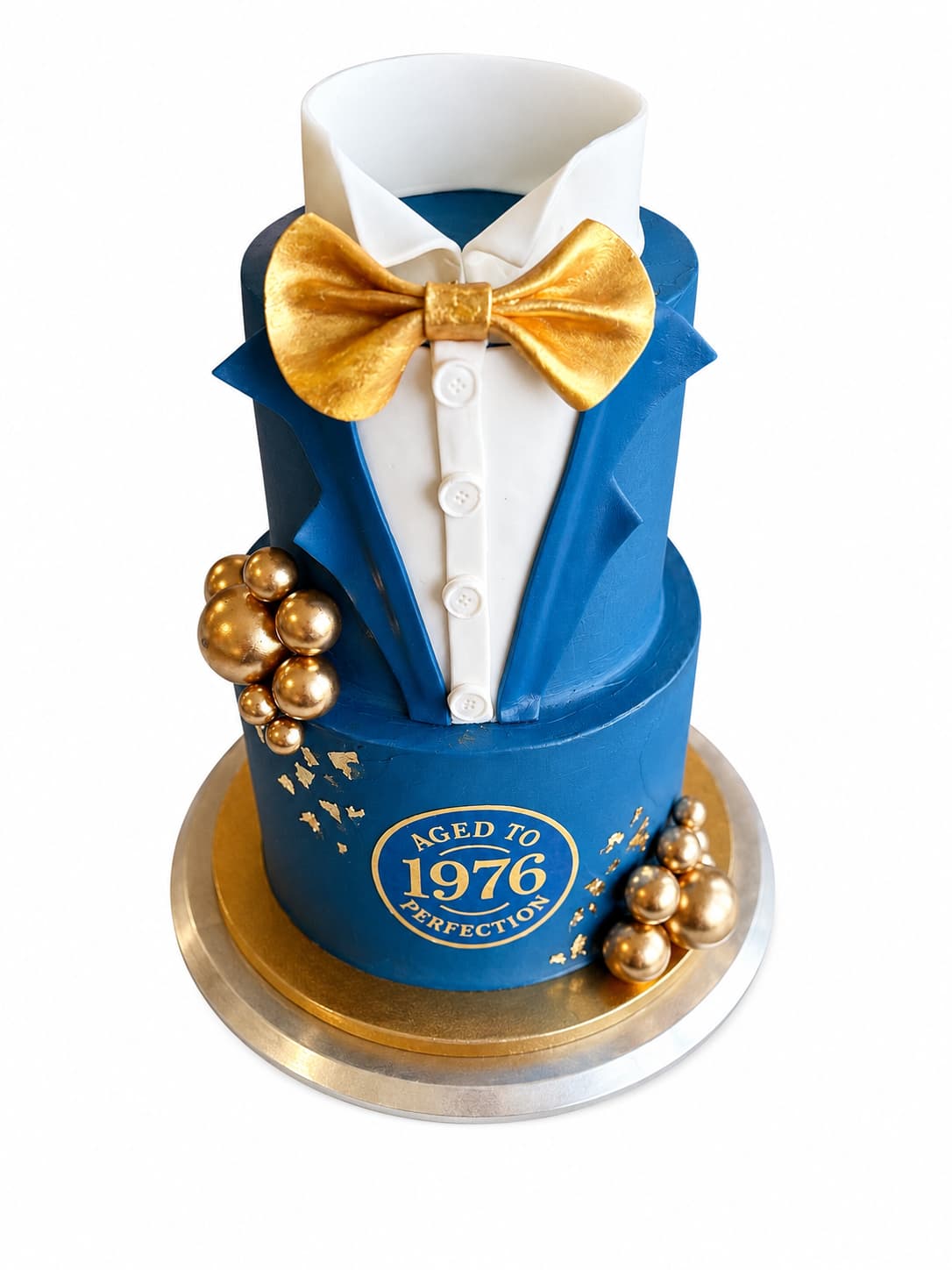 Two-tier tuxedo cake in navy-blue fondant with a white shirt collar and button placket, a large gold bow tie, clusters of gold spheres down the sides, flecks of gold leaf and a round gold monogram reading Aged to 1976 Perfection, on a gold cake board.