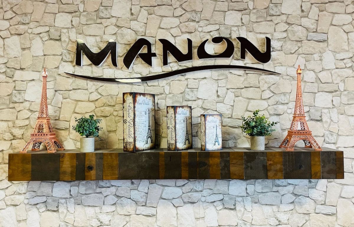 The MANON logo in black metal script mounted on a pale stone wall above a reclaimed-wood mantel, flanked by two copper Eiffel Tower models, two small potted plants and three vintage Paris postcard panels.