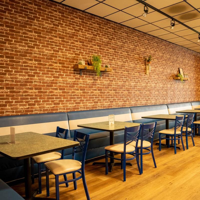 A long navy and cream banquette running the length of an exposed red-brick wall, with square tables, blue caf&eacute; chairs and small wooden shelves of trailing greenery mounted above it.