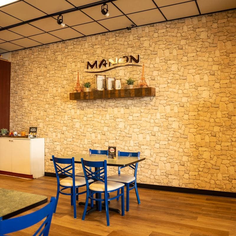 A single four-top table with blue caf&eacute; chairs set against a pale stone wall, beneath a wooden shelf holding the MANON sign, two copper Eiffel Towers and a row of small canisters.