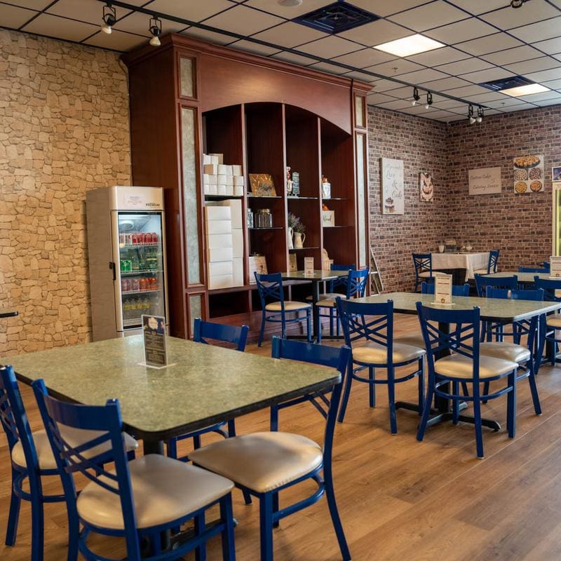 The second half of the dining room, looking past a glass drinks cooler and the wooden hutch toward more tables and blue chairs set along a brick wall.