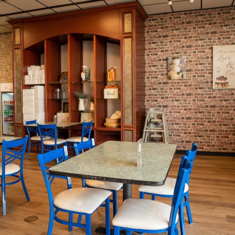 A tall cherry-wood hutch stacked with white pastry boxes, glass jars, a stoneware pitcher and bread baskets, beside a brick wall hung with framed cake prints; a six-top table with blue chairs sits in front of it.
