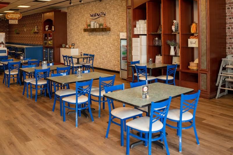 Wide view of the dining room: rows of square tables with blue caf&eacute; chairs on wood-plank flooring, a pale stone feature wall carrying the MANON sign at the back, and a tall cherry-wood hutch on the right.